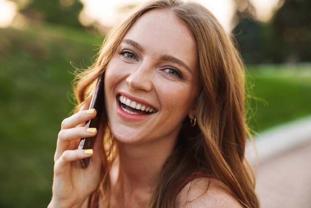 Photo of a laughing positive young ginger woman walking in nature green park outdoors talking by mobile phone.