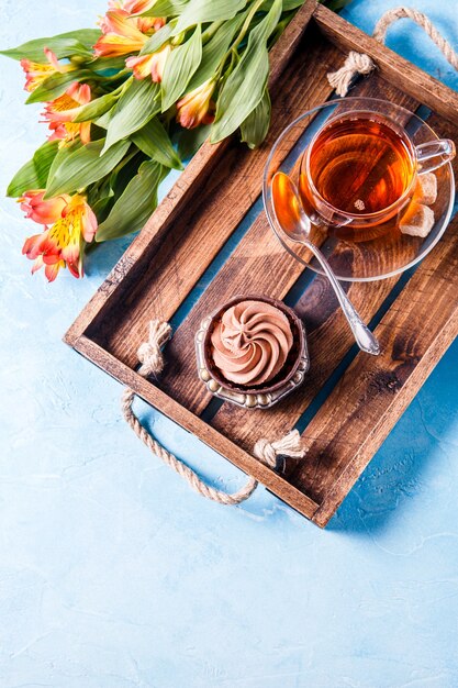 Photo of light breakfast, black tea, cupkake with cream and orange flowers on wooden tray