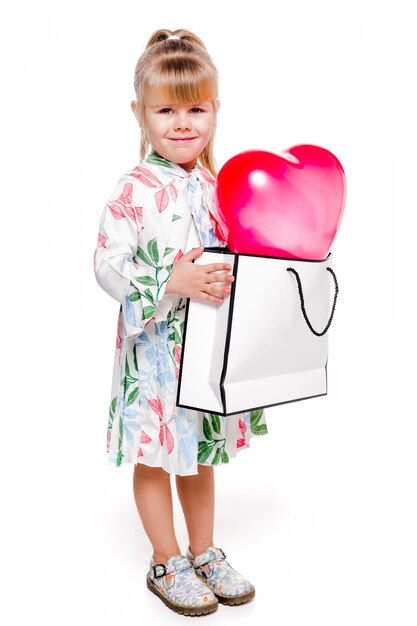 Photo of Little fashionable girl in a white floral dress holds a large bag with a heart-shaped balloon inside.