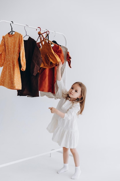 Фотография маленькой девочки, которая выбирает, какую одежду ей надеть среди предложенных на вешалке Фото на белом студийном фоне