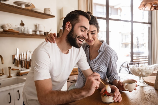 Photo of lovely brunette couple man and woman  drinking coffee and eating desserts during breakfast in kitchen at home