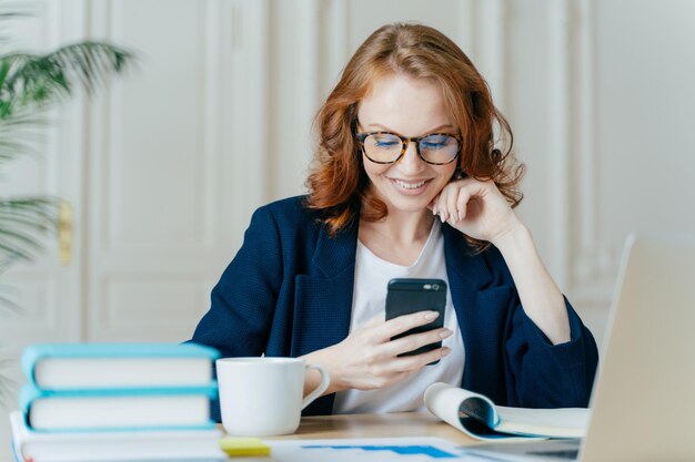 Photo of lovely female sits with smartphone device types feedback works in office on up to date laptop focused into screen of gadget sits at work place with books notepad and hot beverage