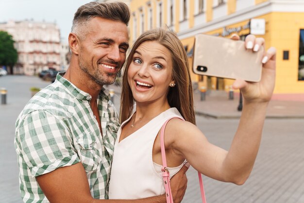Photo of loving young couple in summer clothes smiling and hugging together while taking selfie photo on city street