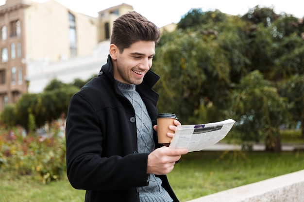 Foto di un macho uomo di 30 anni che indossa una giacca che legge il giornale e beve caffè da asporto mentre cammina per il parco della città