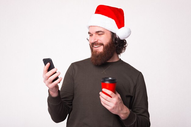 Photo of man with beard wearing xmas hat and using smartphone over white wall