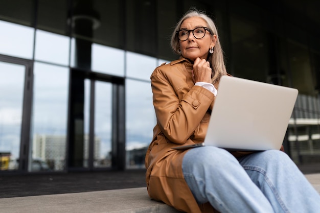 La foto di un'economista femminile adulta matura con capelli grigi in un cappotto marrone si siede con un computer portatile contro