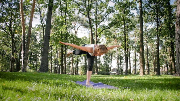 Photo of middle aged smiling woman practicing yoga and meditating at park. Woman stretching and doing fitness on mat at forest