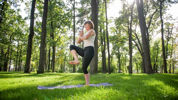 Photo of middle aged woman practicing yoga or fitness on fresh green grass at park. Female physical and mental health. Person in meditation and harmony pf body and soul