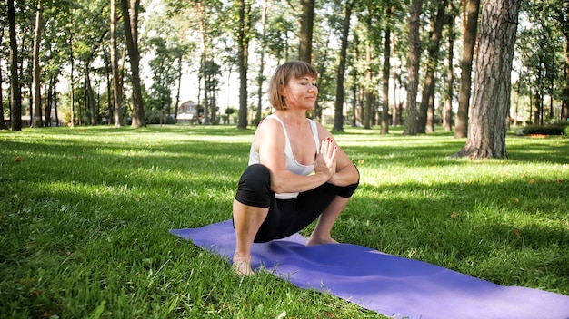 Photo of middle aged woman in sportrs clothes practicing yoga outdoors at park. Middle aged woman stretching and meditating at forest