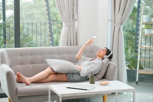 Photo photo of modern asian woman wearing headphones holding cell phone while lying on sofa in bright apartment