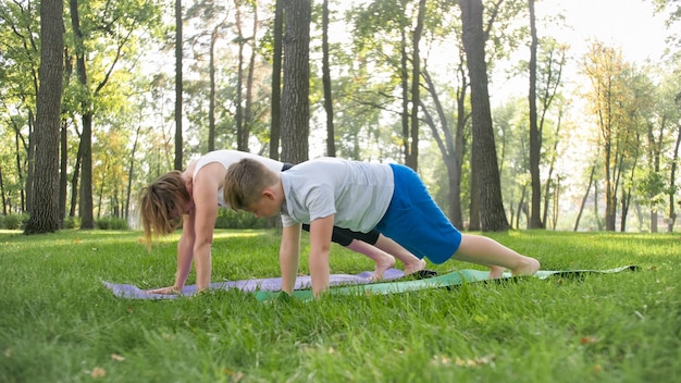 Photo of mother with her teenage boy son practising yoga asana on grass at park. Family doing fitness and sports outtodr at forest