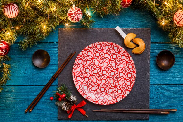 Foto di rami di abete di capodanno, bordo nero, bastoncini di sushi, biscotti di previsione, piatti con motivo rosso su fondo di legno blu