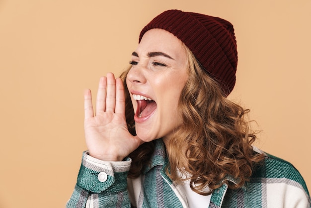 Foto di una bella donna caucasica con un cappello a maglia che urla e guarda da parte isolata sul beige