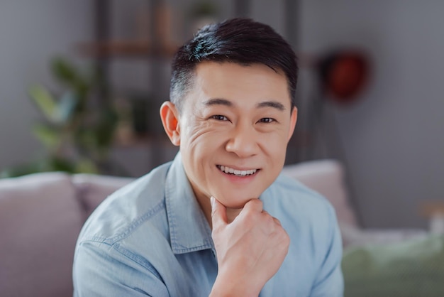 Foto di un uomo giapponese felice e positivo, con un sorriso splendente, il mento del dito che indossa una camicia casual in casa al chiuso