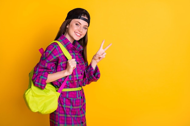 Photo of nice optimistic teen girl show v-sign wear pink dress green bag headwear isolated on yellow color background