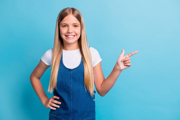 素敵な楽観的なティーンエイジャーのブロンドの長い髪の女の子のポイントの空のスペースの写真は、青い色の背景の上に分離された白いTシャツデニムドレスを着用します