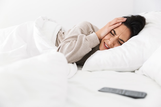 自宅で白いリネンでベッドに横たわっている間、スマートフォンのために頭をつかんでいる不機嫌な女性の写真