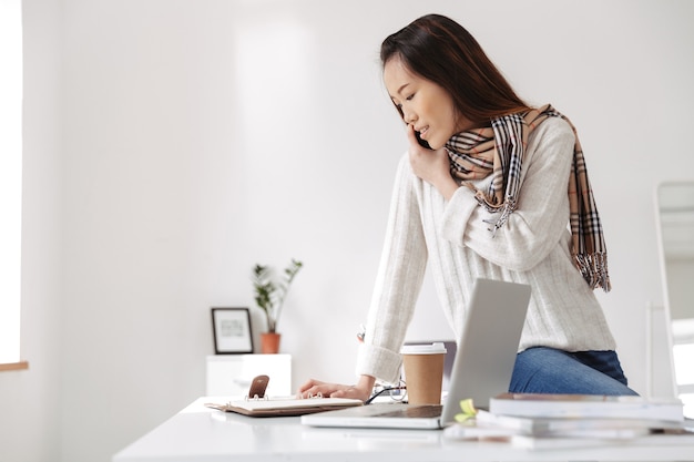 사진 비즈니스 일기를 읽고 사무실에서 테이블에 앉아있는 동안 웃고 흰색 스웨터를 입고 낙관적 아시아 여성 노동자의 사진
