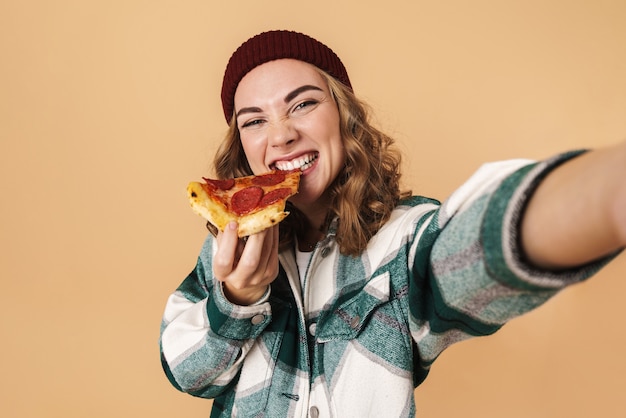Фото Фото довольно счастливой женщины в вязаной шляпе, делающей селфи и едящей пиццу, изолированную на бежевом