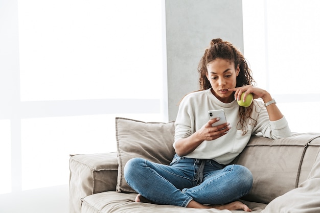 Фотография довольной афро-американской женщины, использующей мобильный телефон, сидя на диване в светлой квартире