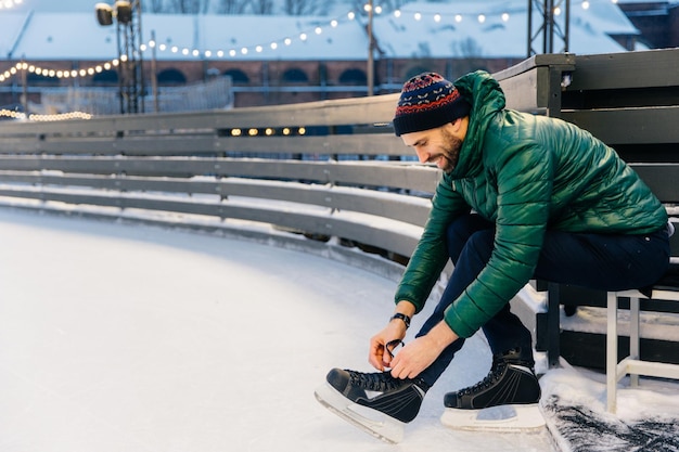 写真 幸せな表情で熟練した男性モデルの写真は、緑のアノラックに身を包んだスケート靴をひもで結び、氷のリングに座って、好きな趣味に携わる予定です。男性は屋外で楽しくエンターテイメントを楽しんでいます。