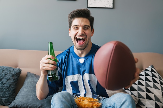 写真 リビングルームでスポーツの試合を見ながら、ラグビーボールの叫び声とビールを飲んで喜んでいる若い男の写真