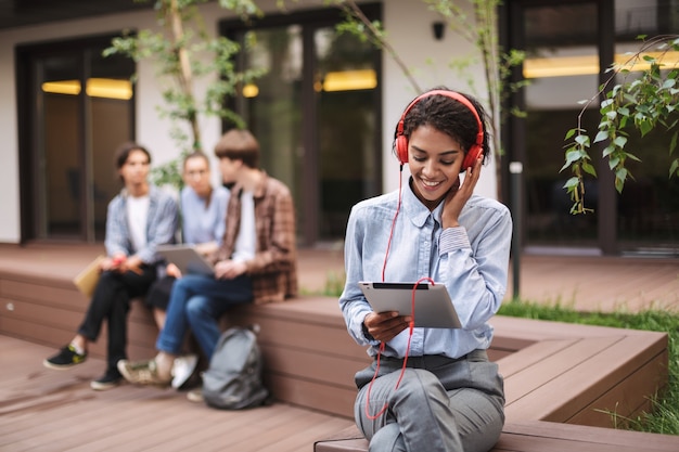 写真 大学の中庭で時間を過ごしている間、赤いヘッドフォンとタブレットを手にベンチに座っている若い女性の写真