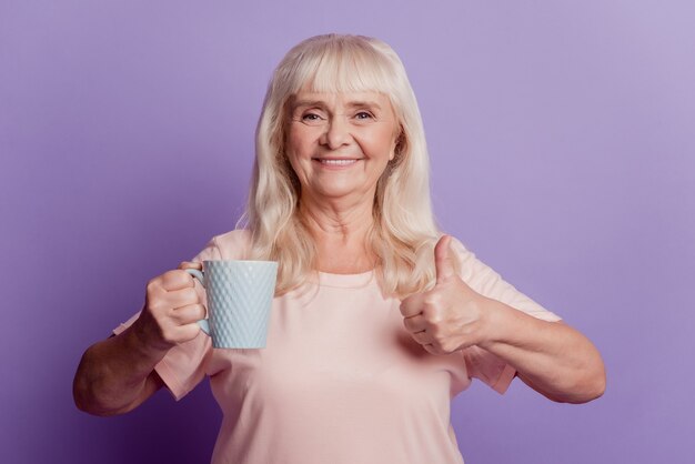 Фотография старой женщины, держащей чай, кофейную чашку, показывает знак большого пальца на фиолетовом фоне