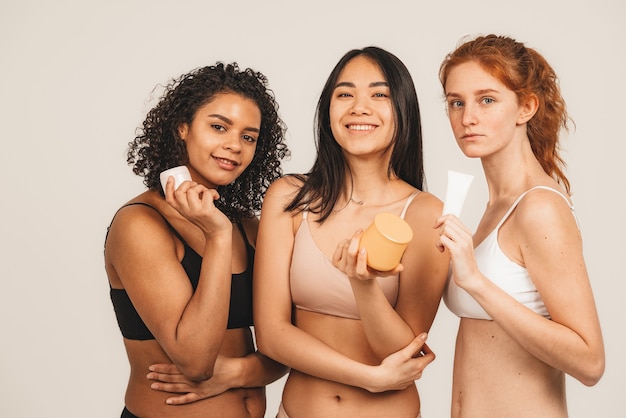 Photo photo of optimistic cheery young women, multiracial friends wear underwear, hold cream and beauty products