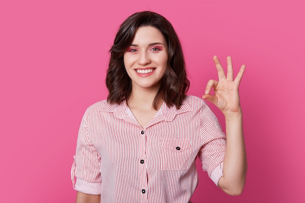 Photo of optimistic joyful beautiful woman has professional makeup, wavy dark hair, makes okay gesture, wears striped shirt, isolated over pink