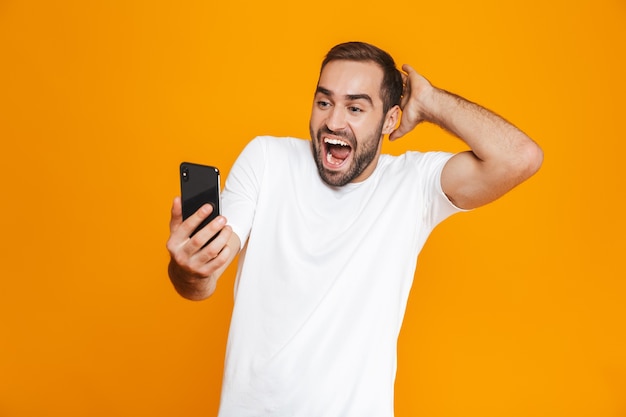 Photo of optimistic man 30s in casual wear smiling and holding smartphone, isolated