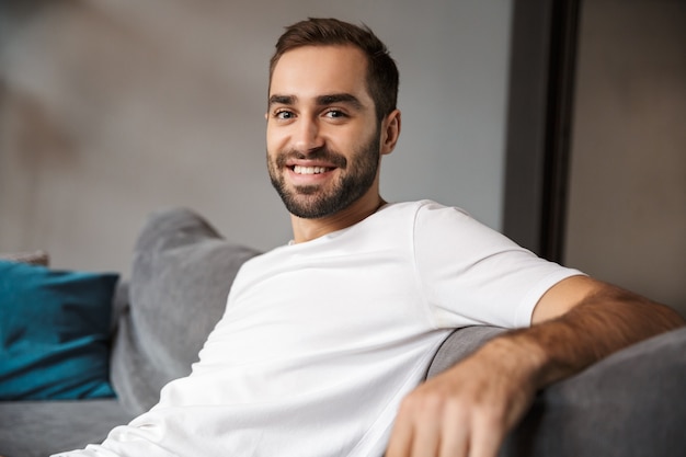 Photo of optimistic man 30s wearing casual t-shirt smiling while sitting on sofa in living room