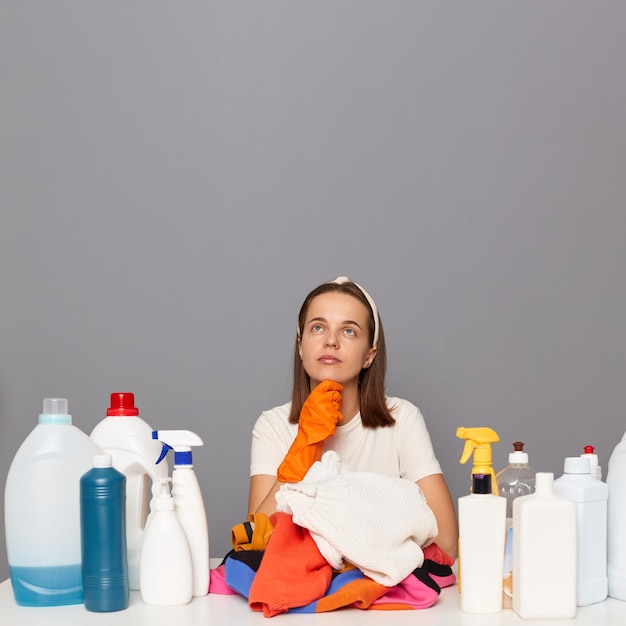 Photo of pensive Caucasian woman wears rubber protective gloves and headband surrounded with bottles of detergents for laundry looking up at copy space for advertisement