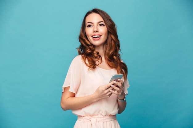 Photo of pleased amazing woman holding cell phone and looking aside on copyspace, isolated over blue wall