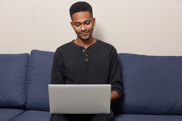 Photo of pleased dark skinned guy has beard, wears black sweater, poses at comfortable sofa, installs application