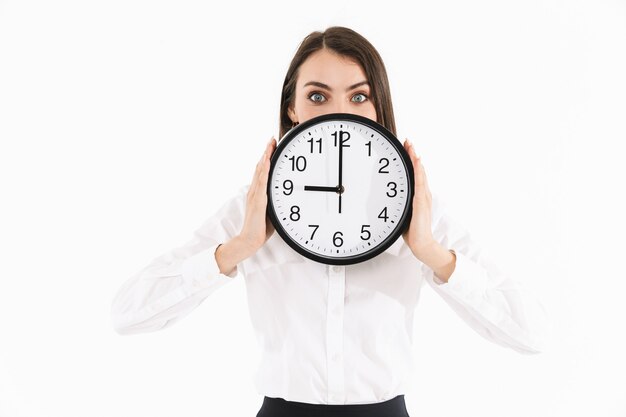 Foto di una donna d'affari soddisfatta lavoratrice vestita con abiti formali che tiene in mano un grande orologio da parete mentre lavora in ufficio isolato su un muro bianco