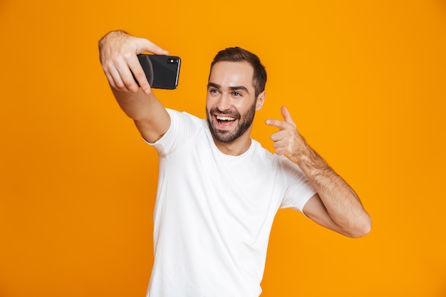 Photo of pleased guy 30s in casual wear laughing and taking selfie on cell phone, isolated