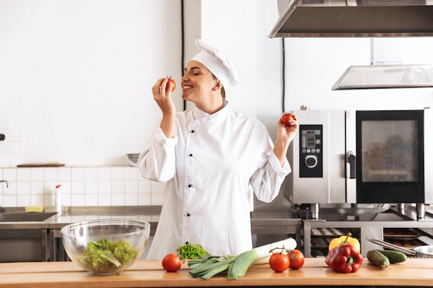 Photo of pleased woman chef wearing white uniform cooking meal with fresh vegetables, in kitchen at the restaurant