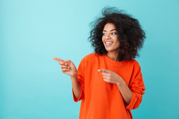 Photo of pleased woman in red shirt looking aside and pointing fingers on copyspace, isolated over blue wall