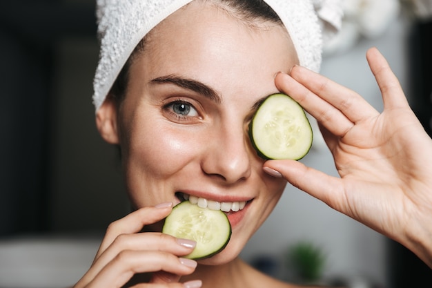 Photo of pleased woman with towel on head holding cucumber slices at her face