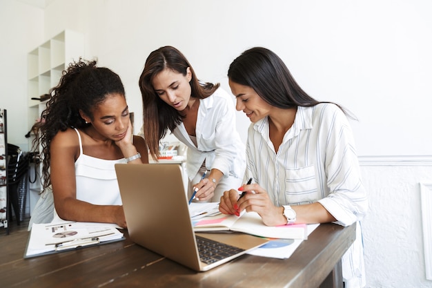 Фотография довольных молодых деловых женщин коллег в офисе в помещении с помощью портативного компьютера, разговаривая друг с другом.
