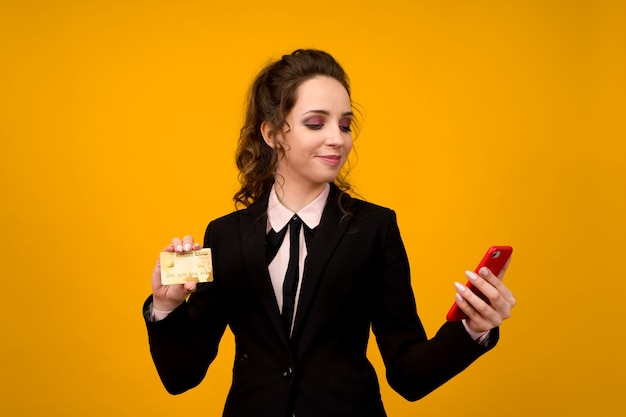 Photo of pleased young woman posing isolated over yellow wall background using mobile phone holding debit card