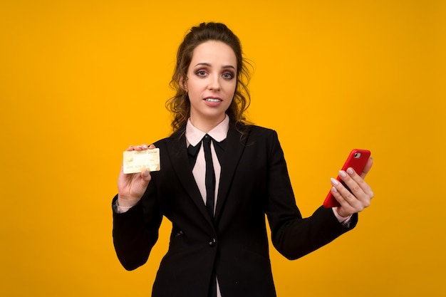 Photo of pleased young woman posing isolated over yellow wall background using mobile phone holding debit card.