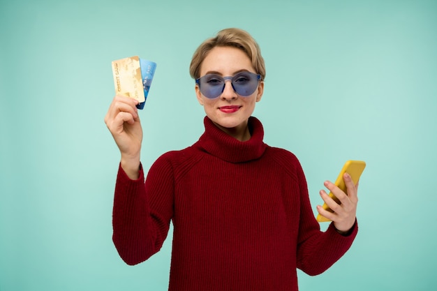 Foto di felice giovane donna con acne problema viso pelle in posa isolato su sfondo blu muro utilizzando il telefono cellulare tenendo la carta di debito.