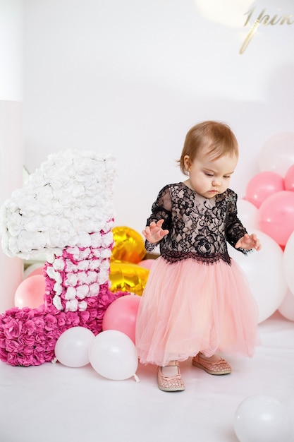 Photo portrait of a birthday girl 1 year old in a pink dress with pink balloons. The child at the holiday smiles, children's emotions. Birthday party