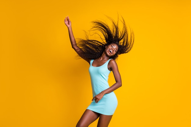 Photo portrait of cute active african american girl dancing with one hand in air hair flying wearing blue mini dress isolated on vivid yellow colored background