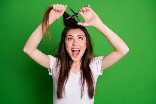 Photo portrait of excited woman with open mouth cutting hair isolated on vivid green colored background