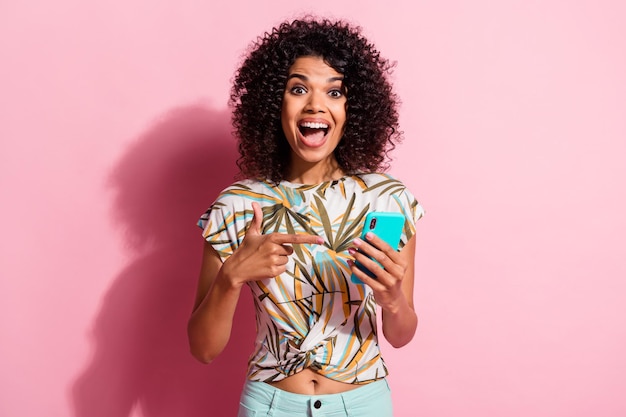 Photo portrait of excited woman with open mouth holding phone in one hand pointing finger at it isolated on pastel pink colored background