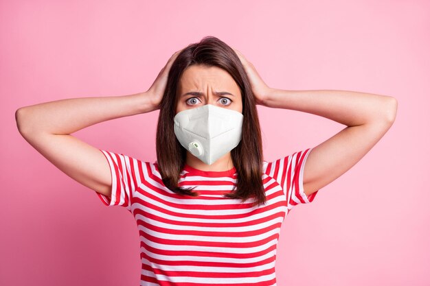 Photo portrait of frightened brunette holding head with hands wearing white respiratory mask isolated on pastel pink colored background