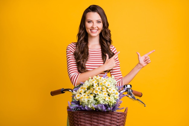 Foto ritratto di ragazza che punta con le dita su copyspace in bicicletta con cesto di fiori isolato su sfondo di colore giallo brillante